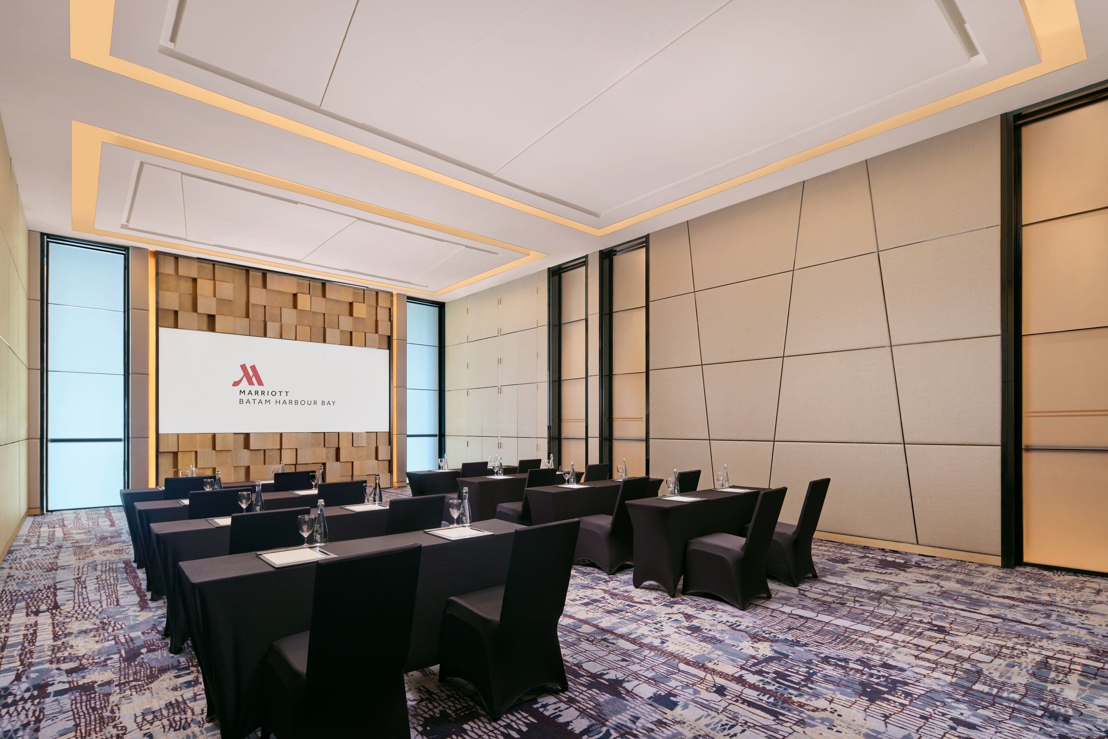 Un salón de conferencias con una pantalla grande mostrando el logotipo de Marriott, varias mesas con sillas y una alfombra estampada.
