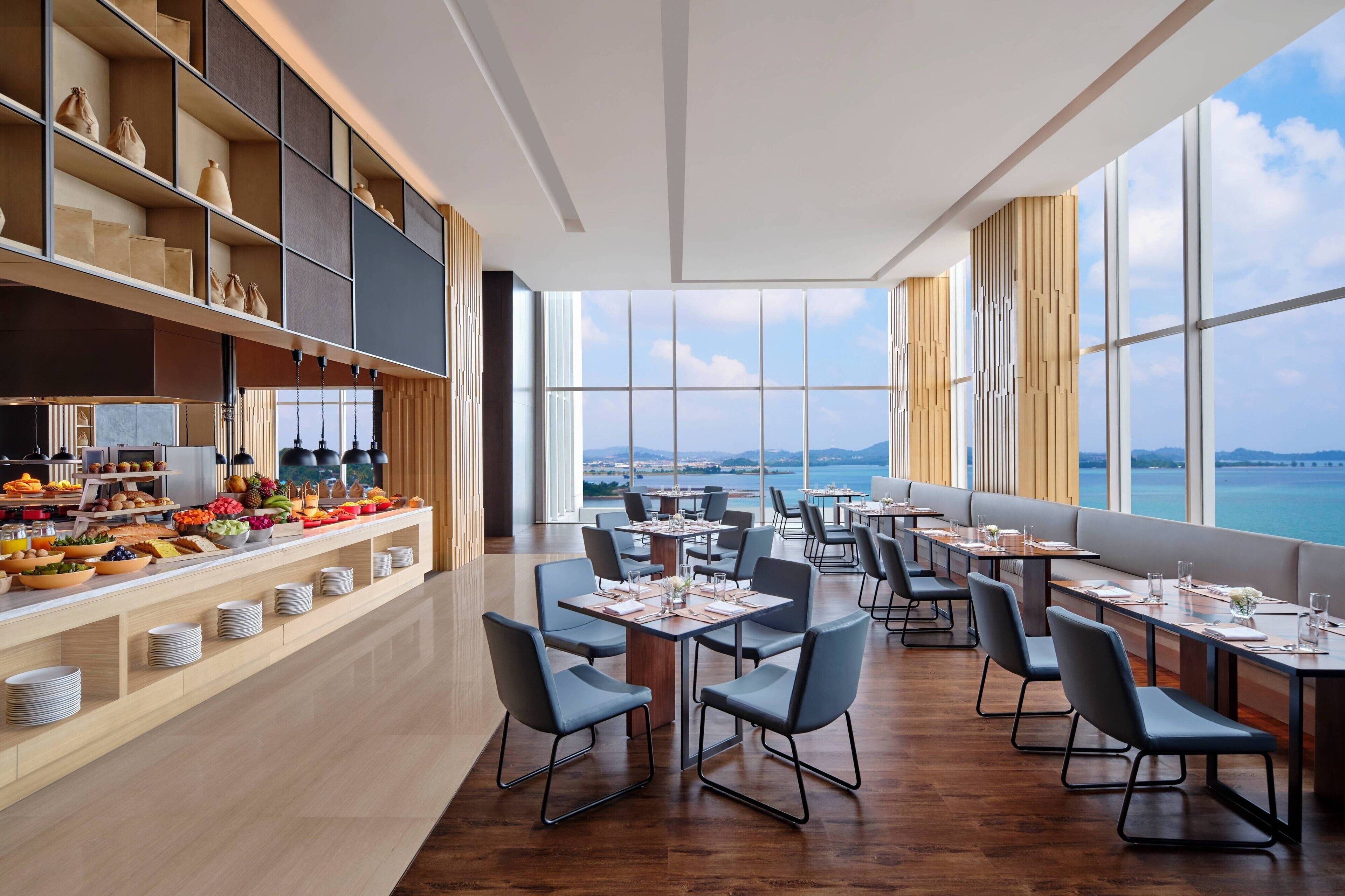 A modern restaurant with a buffet on the left, large windows offering a view of the sea, and neatly arranged tables and chairs.