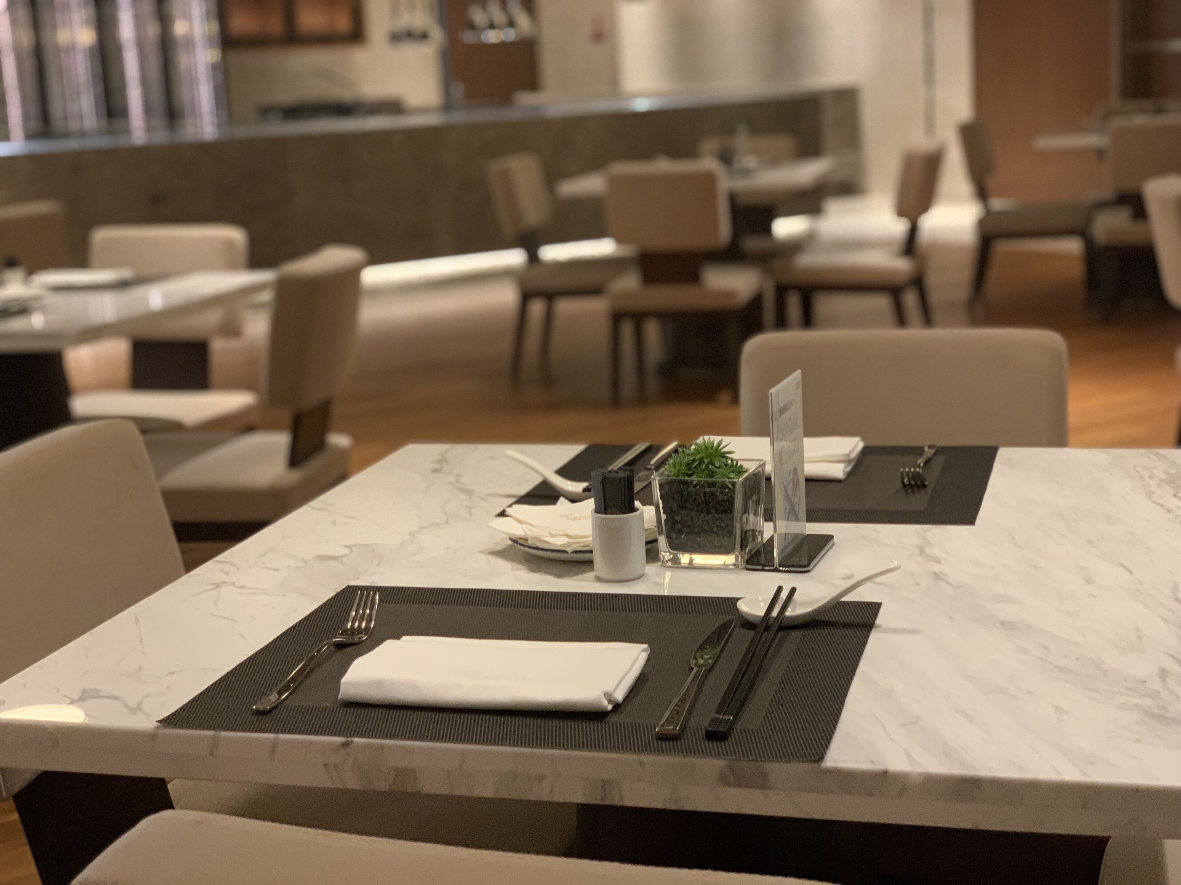 A dining area with tables set for two, featuring a marble top, black placemats, and a small potted plant.