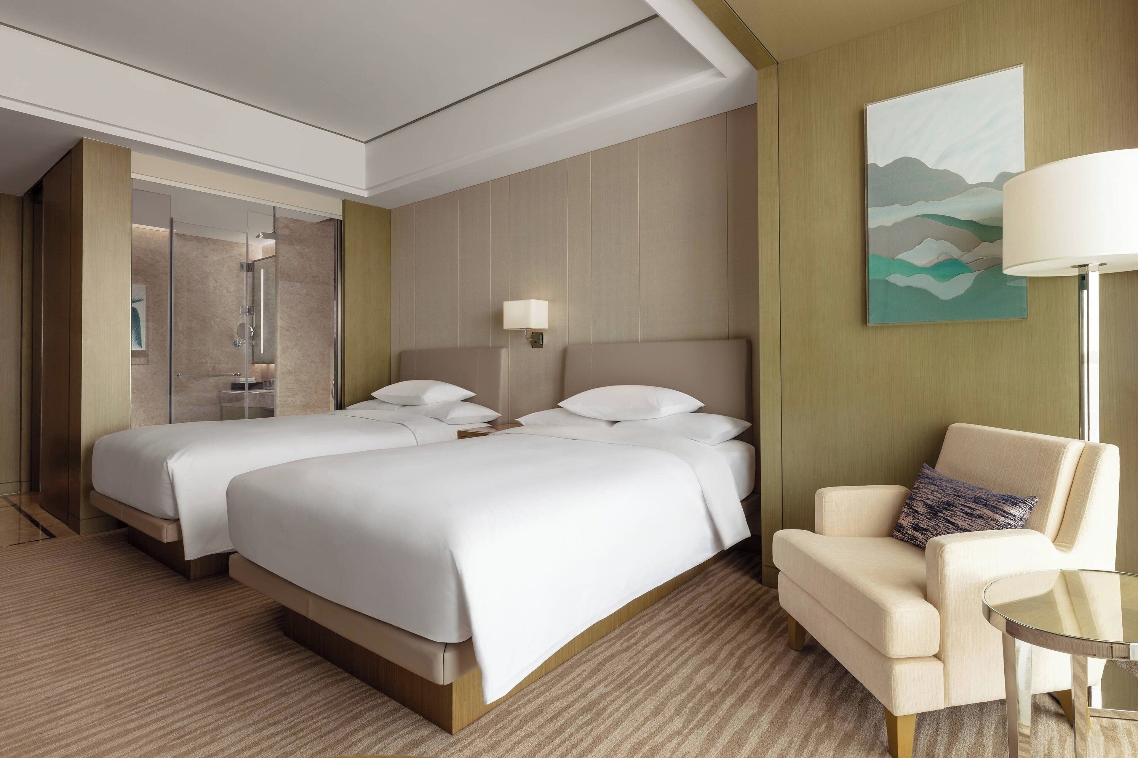 A hotel room with two beds, a sitting area with a chair and a table, a painting on the wall, and a lamp.