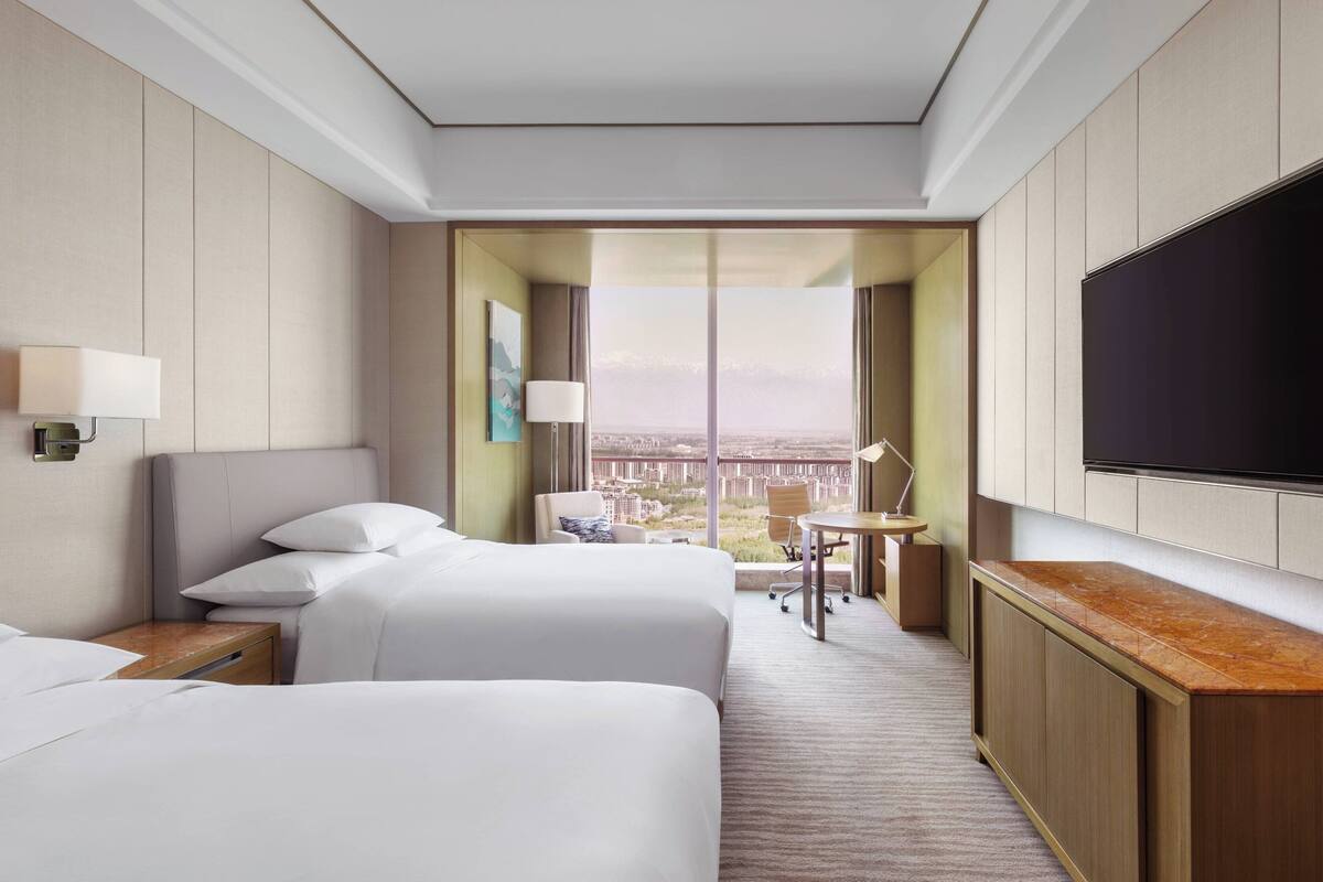 A hotel room with two beds, a TV, a desk, and a view of a cityscape.