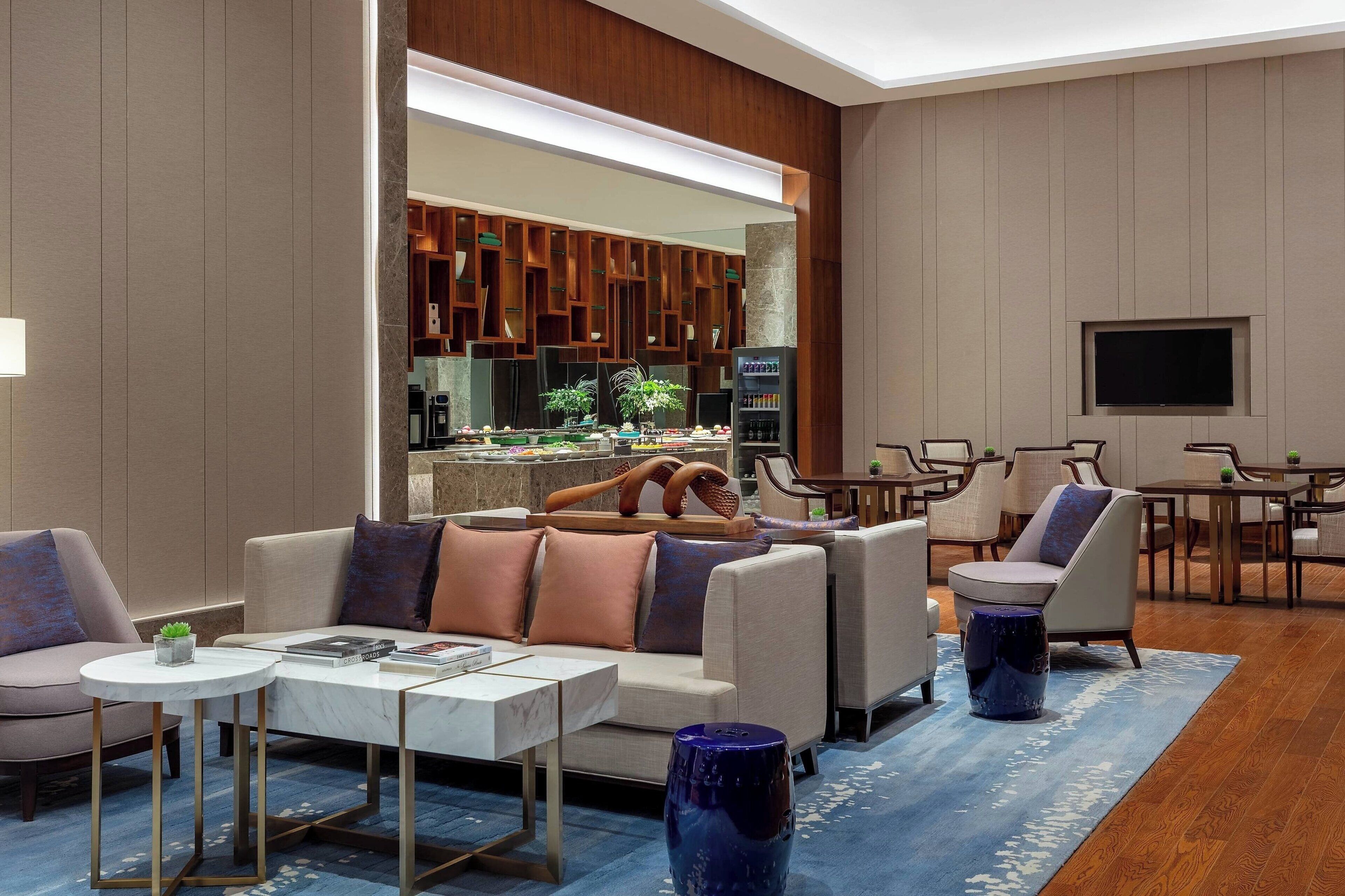 Un salon moderne avec un canapé, une table basse et des fauteuils. Un téléviseur est fixé au mur, et on aperçoit un coin cuisine à travers une porte ouverte.