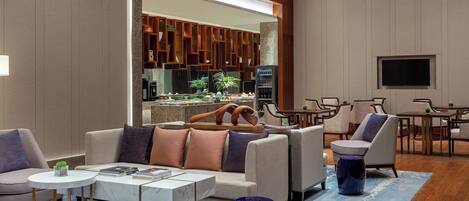 Un salon moderne avec un canapé, une table basse et des fauteuils. Un téléviseur est fixé au mur, et on aperçoit un coin cuisine à travers une porte ouverte.