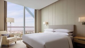 Habitación de hotel con una cama grande, una silla, una mesita de noche y vistas a la ciudad a través de amplias ventanas.