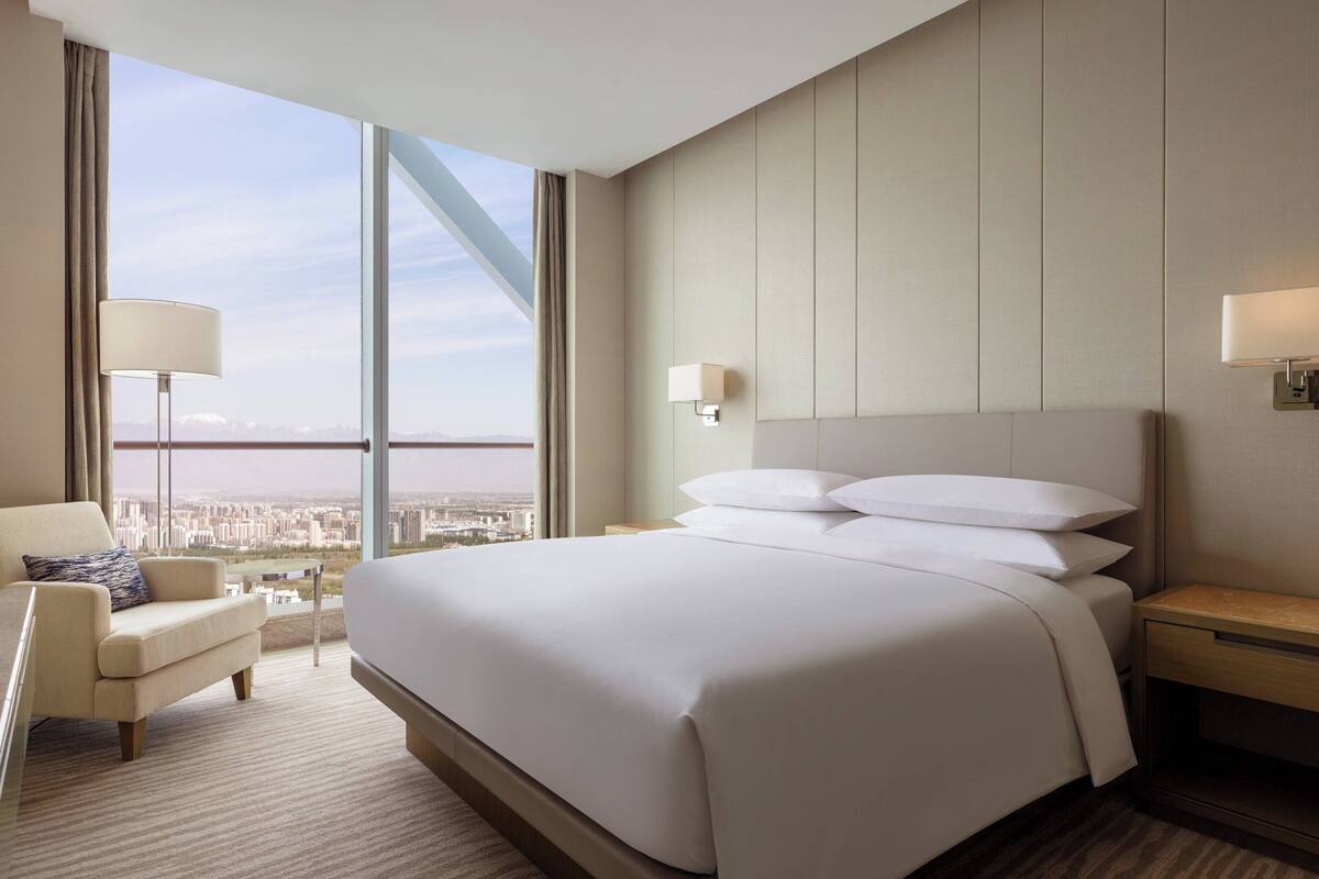 A hotel room with a large bed, a chair, a nightstand, and a view of the city through large windows.