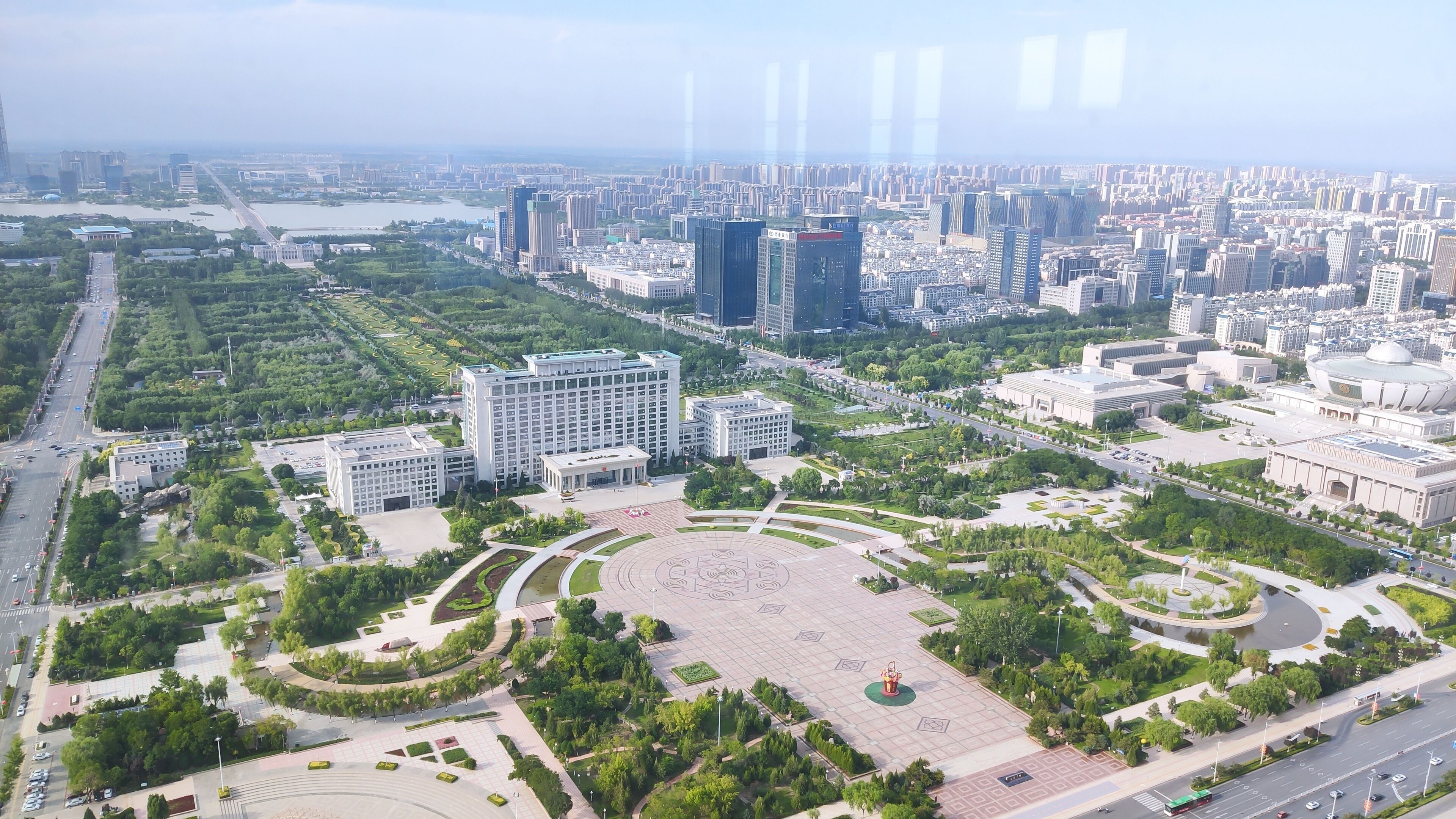 An aerial view of a cityscape with a large central plaza surrounded by modern buildings, roads, and greenery.