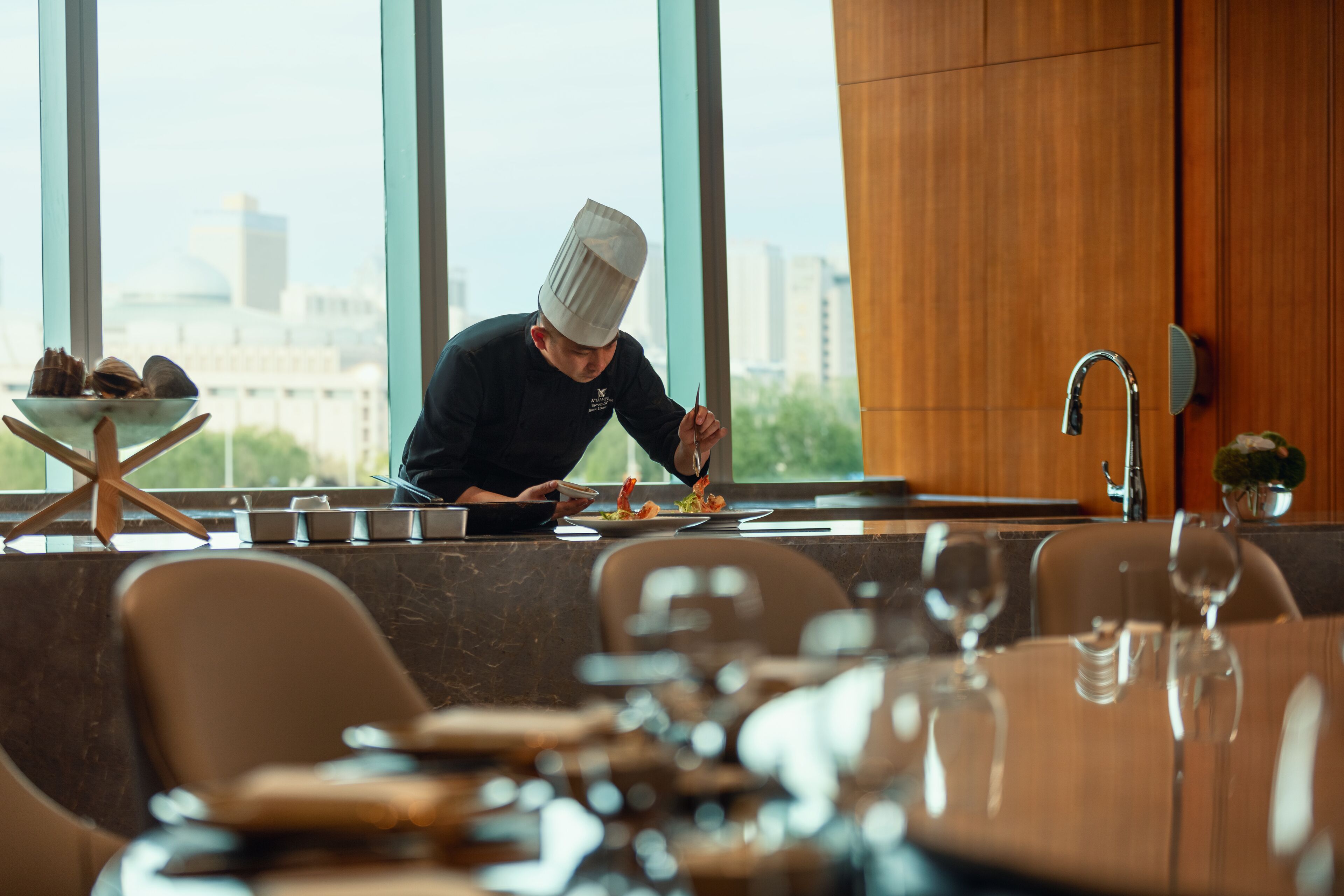 A chef preparing food in a modern kitchen with large windows, a view of the city, and a dining area with tables and chairs.
