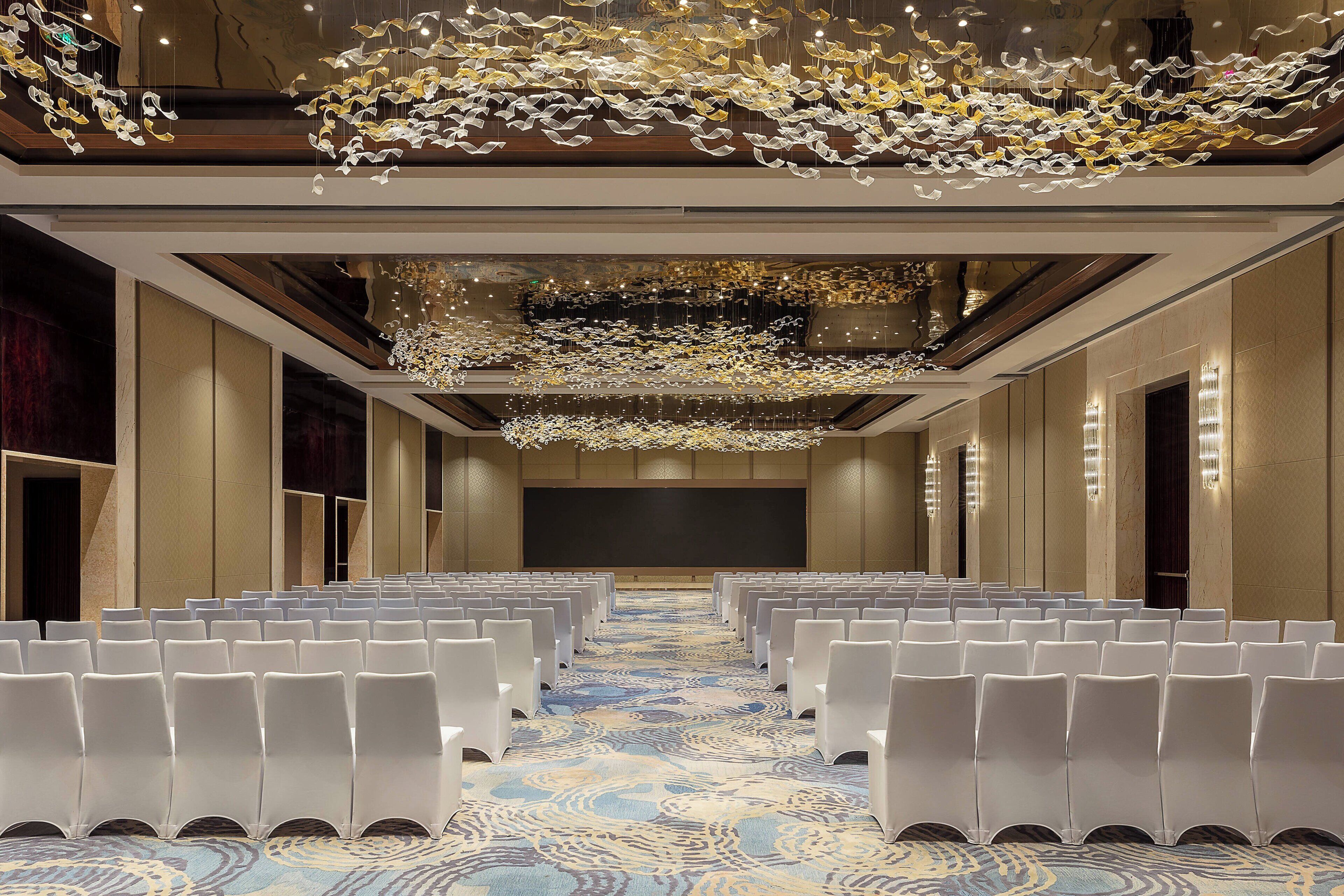 A conference room with rows of white chairs, a patterned carpet, and a ceiling with hanging light fixtures.