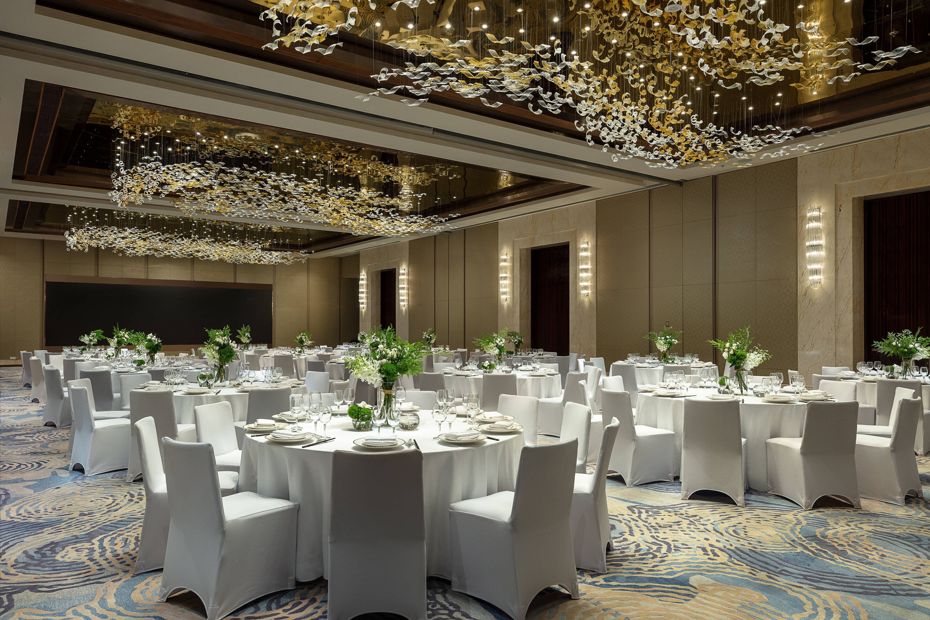 A banquet hall with round tables set for dining, white chairs, and a decorative chandelier.