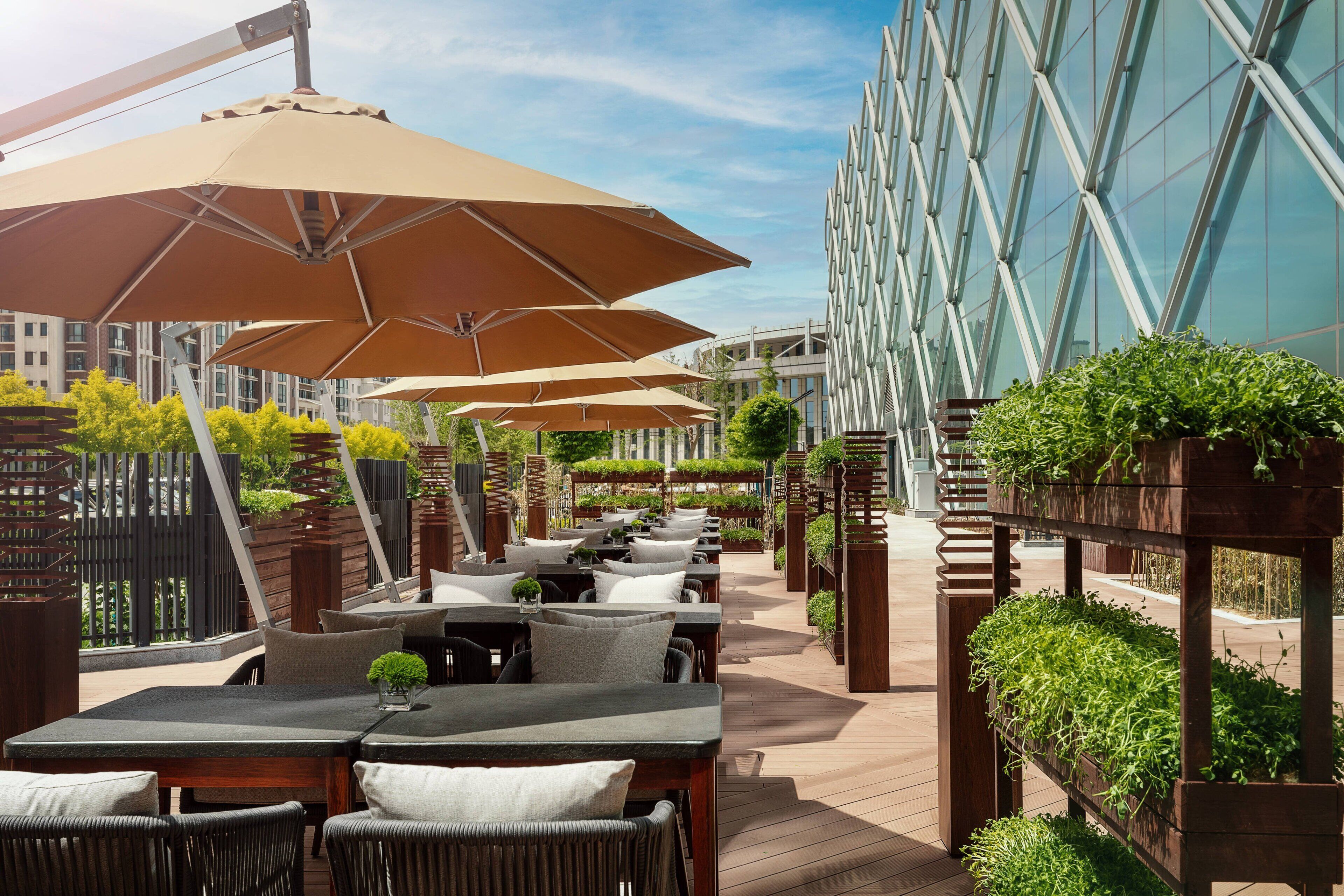 Een buitenterras met meerdere tafels en stoelen, grote parasols, en een modern gebouw met glazen ramen op de achtergrond.