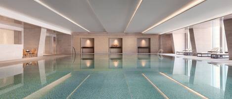 A spacious indoor swimming pool with clear blue tiles, recessed lighting, and seating areas along the sides.