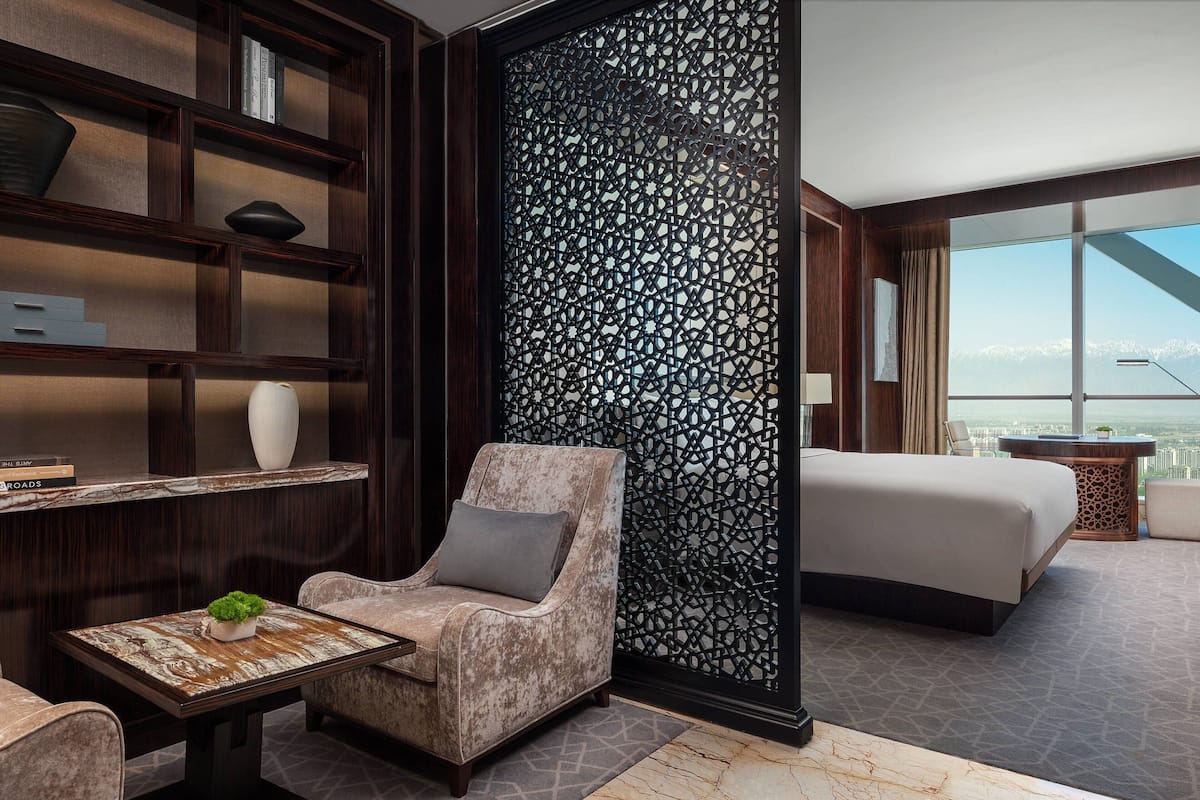 Une chambre d’hôtel moderne dotée d’un grand lit, d’un coin salon avec un fauteuil et une table, d’un paravent décoratif et offrant une vue sur les montagnes.