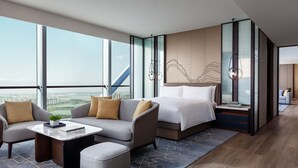 Una habitación de hotel moderna con un ventanal grande, un sofá, una cama, una mesita pequeña y una mesita de noche.