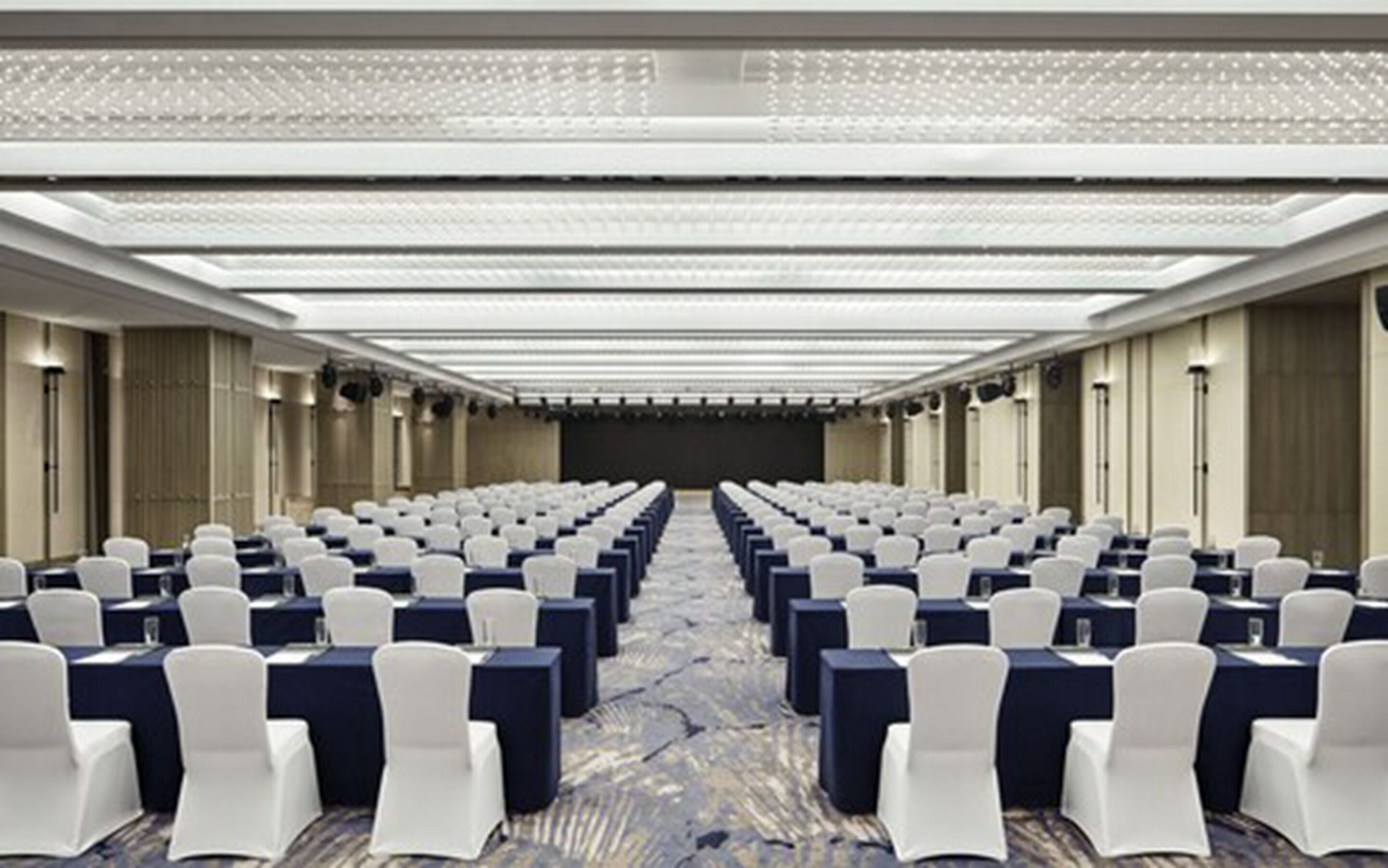 A conference room with rows of white chairs and tables, a patterned carpet, and a ceiling with recessed lighting.