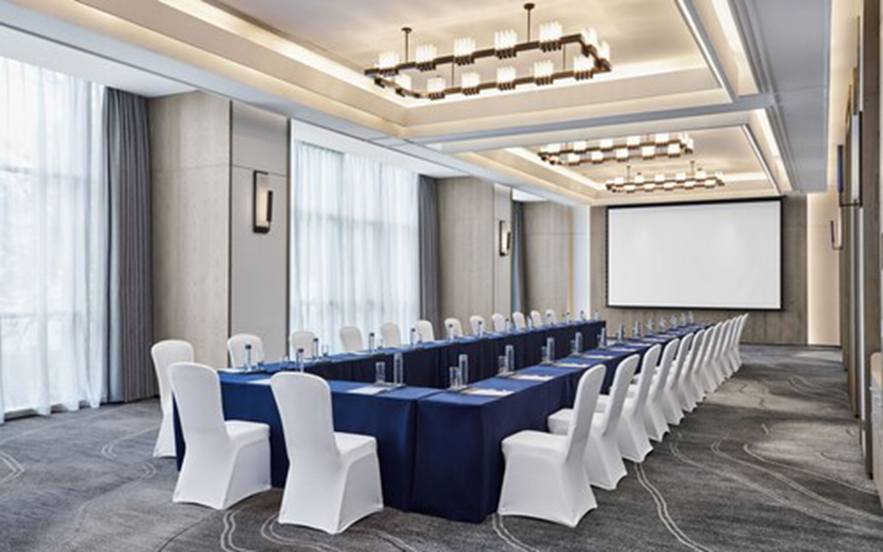 Un salón de conferencias con una larga mesa, sillas blancas y una pantalla para proyector.