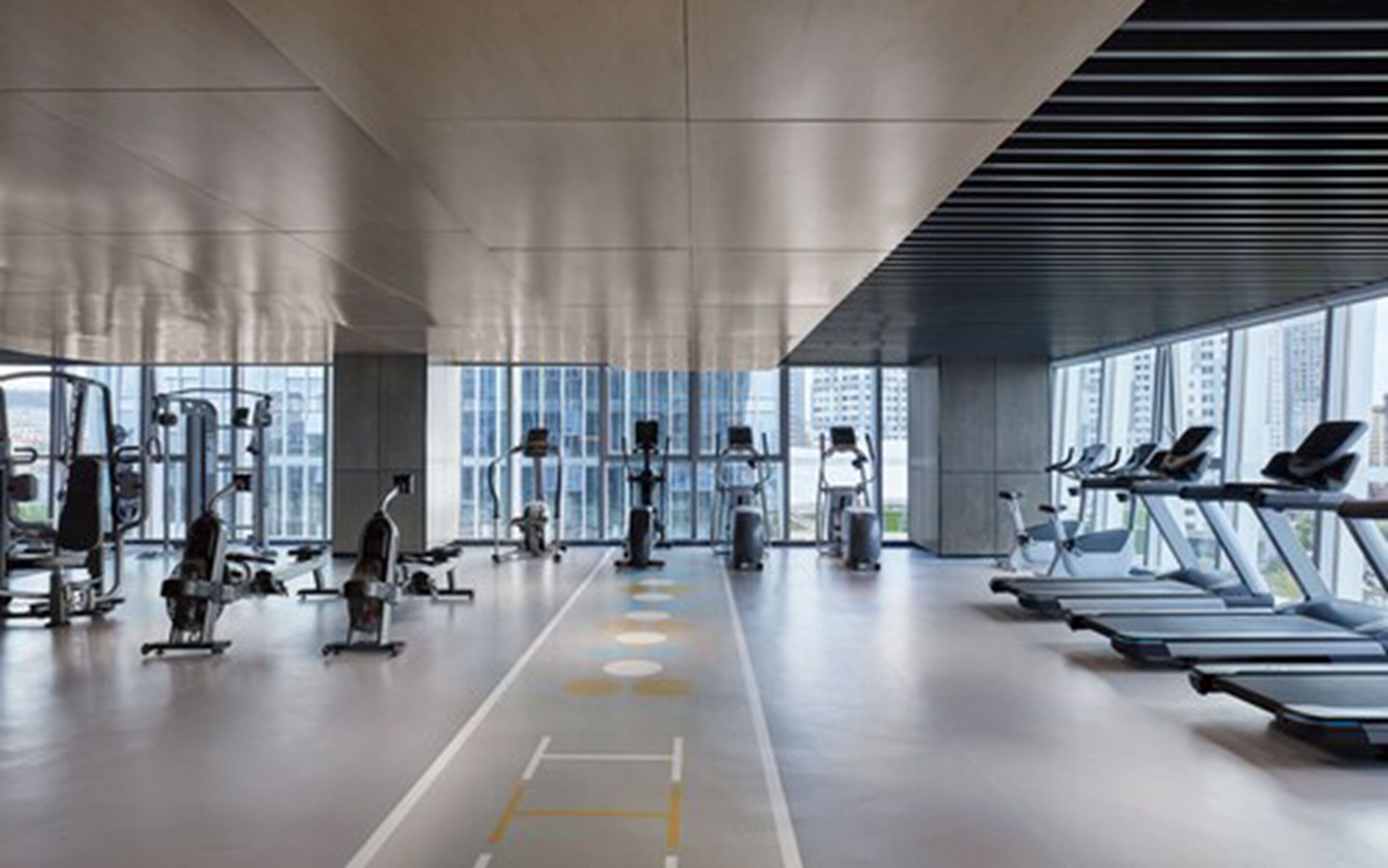 A modern gym with cardio machines, weight stations, and a large window offering a city view.