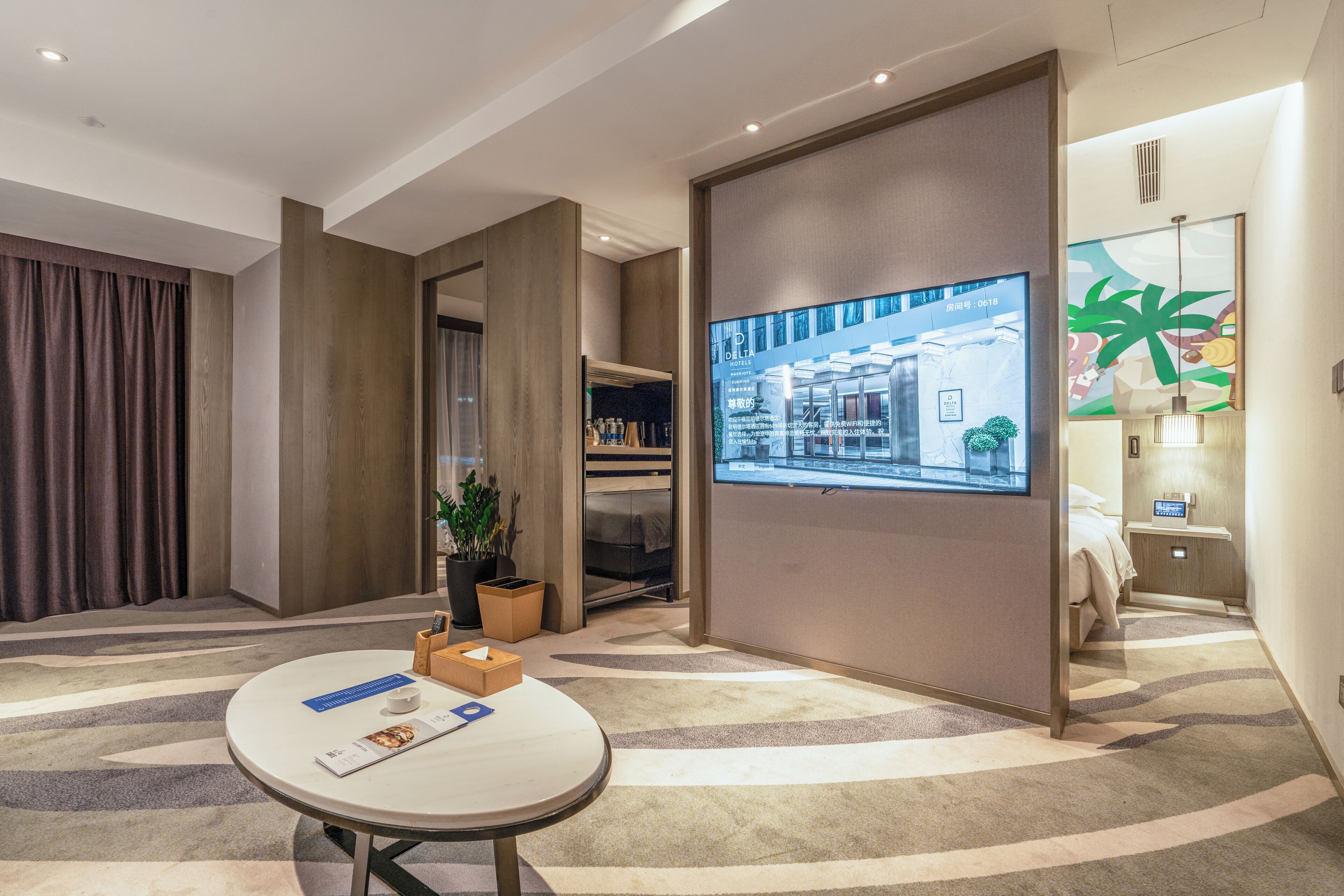 A hotel room with a large TV screen, a round table with a magazine, a plant, and a view of the hotel lobby.