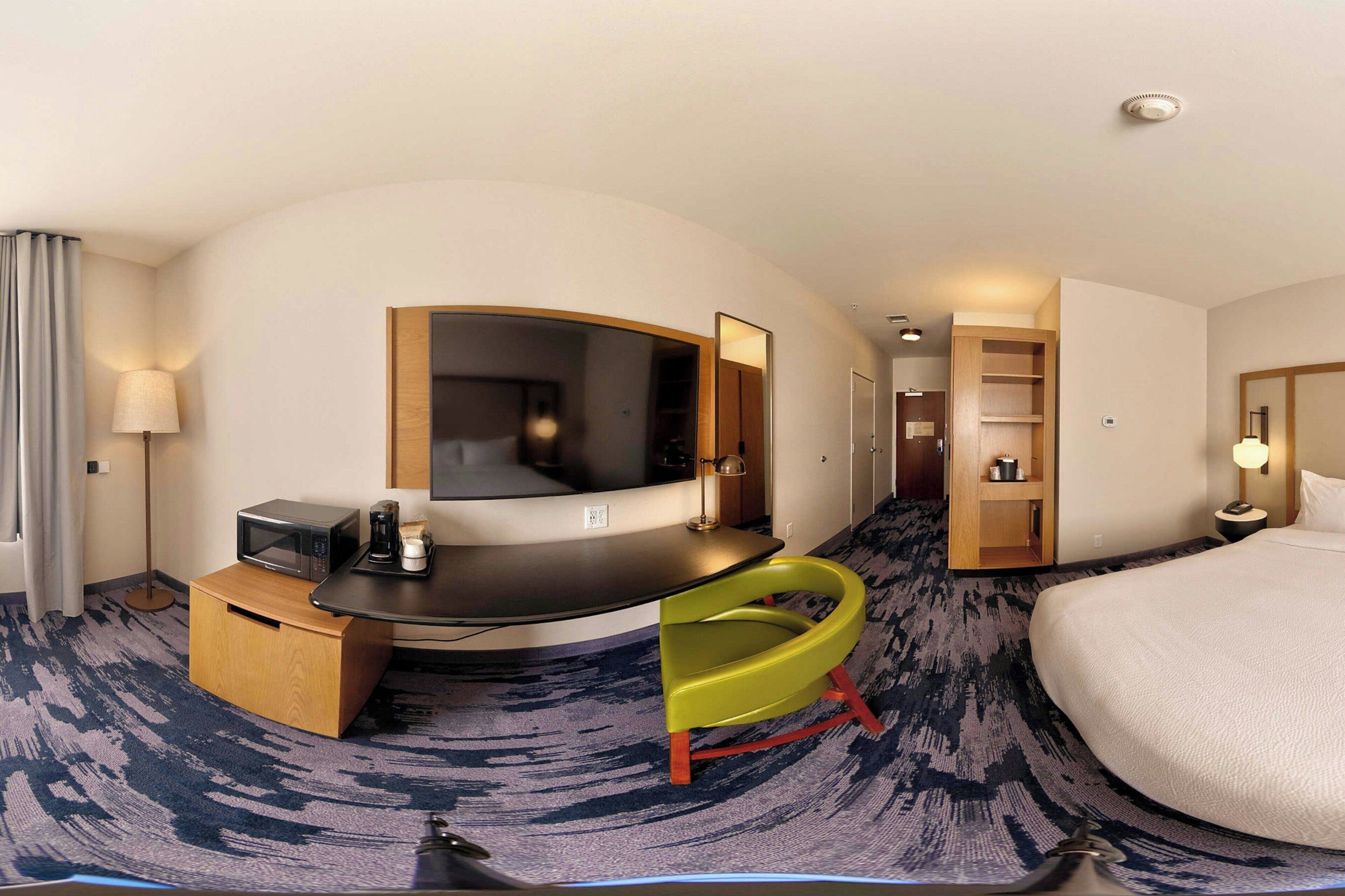 Ein Hotelzimmer mit einem gebogenen Bett, einem Schreibtisch mit Mikrowelle, einem Sessel, einem Spiegel, einem Kleiderschrank und einem kleinen Regal mit Gegenständen.