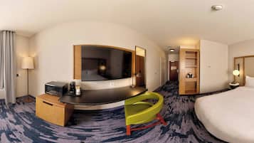 Ein Hotelzimmer mit einem gebogenen Bett, einem Schreibtisch mit Mikrowelle, einem Sessel, einem Spiegel, einem Kleiderschrank und einem kleinen Regal mit Gegenständen.