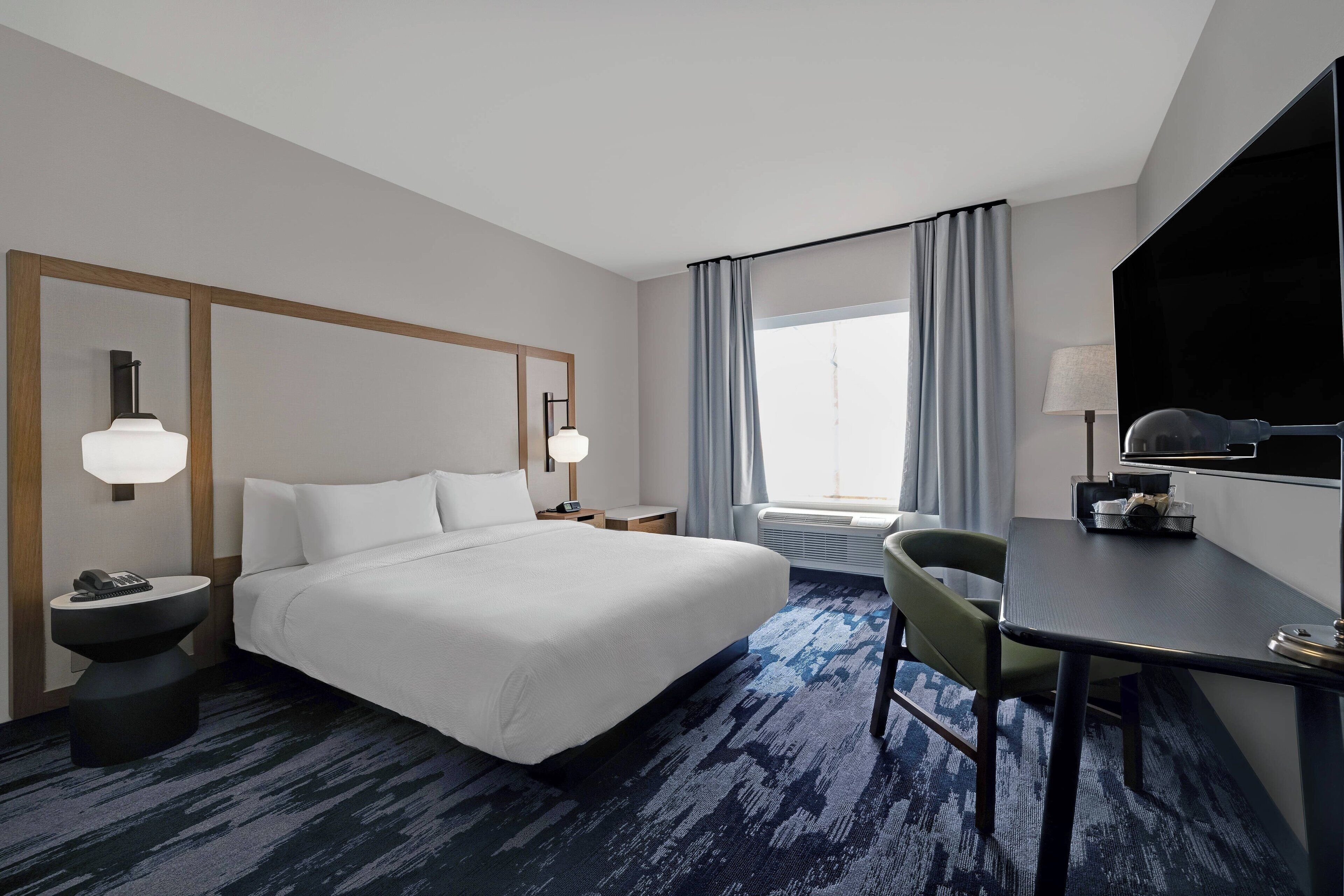 A hotel room with a large bed, a desk, a chair, a TV, and a window with curtains.