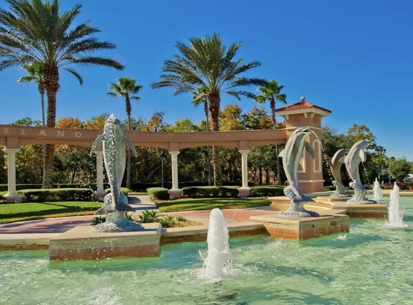 Ein Springbrunnen mit mehreren Wasserstrahlen und Statuen, umgeben von Palmen und wolkenlosem, blauem Himmel.