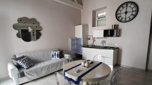 A modern living room with a sofa, a dining table with chairs, a kitchen area with a refrigerator, and a wall clock. - Casa Bolle (Sicilia)