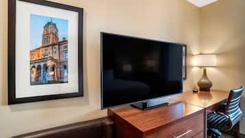 Habitación de hotel con una televisión de pantalla plana grande sobre un escritorio de madera, un cuadro enmarcado de un edificio en la pared, una lámpara de escritorio y un sillón de cuero.