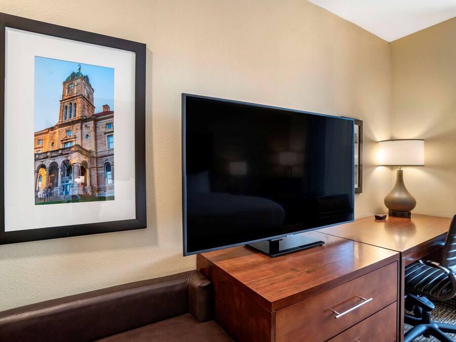 Habitación de hotel con una televisión de pantalla plana grande sobre un escritorio de madera, un cuadro enmarcado de un edificio en la pared, una lámpara de escritorio y un sillón de cuero.