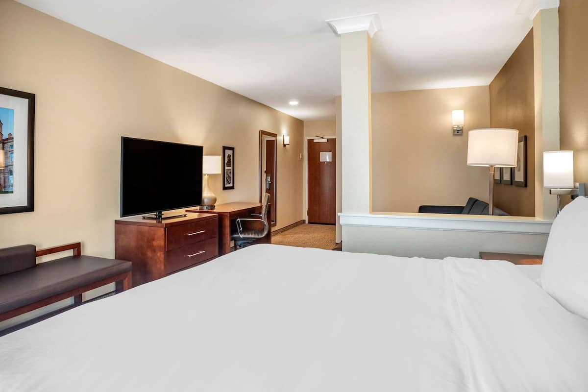 Camera d'albergo con un letto grande, una TV a schermo piatto, una scrivania con una sedia, uno specchio e una porta che dà su un'altra stanza.