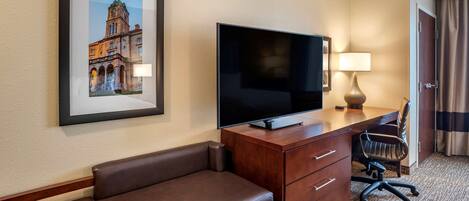 Habitación de hotel con televisor de pantalla plana grande, escritorio de madera, banca de cuero, una silla y un cuadro en la pared.