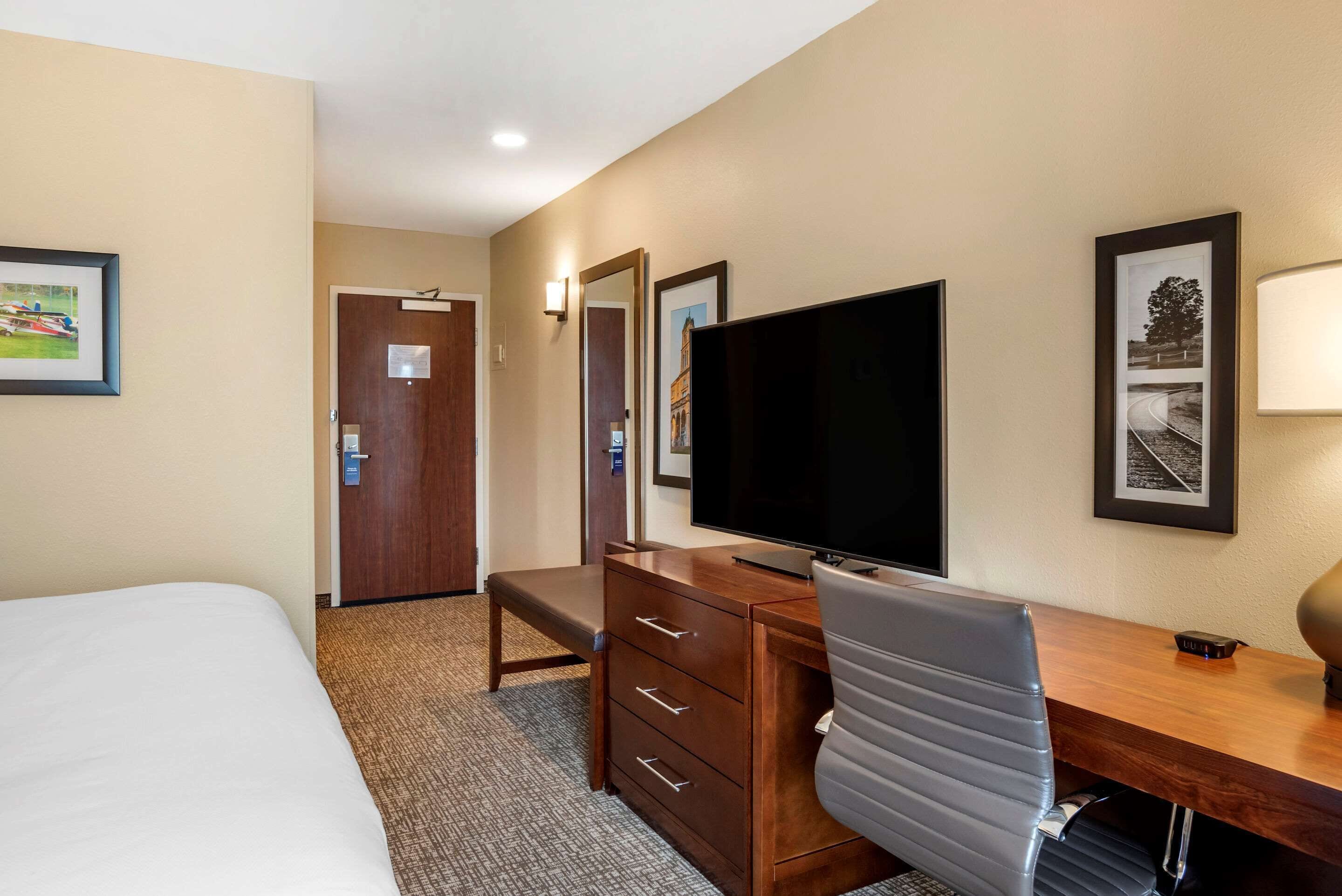 A hotel room with a bed, a desk with a chair, a flat-screen TV, a wooden dresser, and framed pictures on the walls.