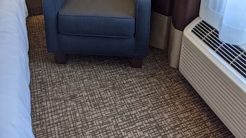 Hotellihuone, jossa on sininen nojatuoli, yöpöytä lampun kanssa, sänky ja ikkuna, jossa on verhot.