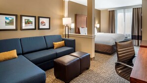 A hotel room with a blue sofa, a brown ottoman, a bed, and a desk with a chair. There are framed pictures on the wall and a window with curtains. - Comfort Inn & Suites Harrisonburg (Harrisonburg)
