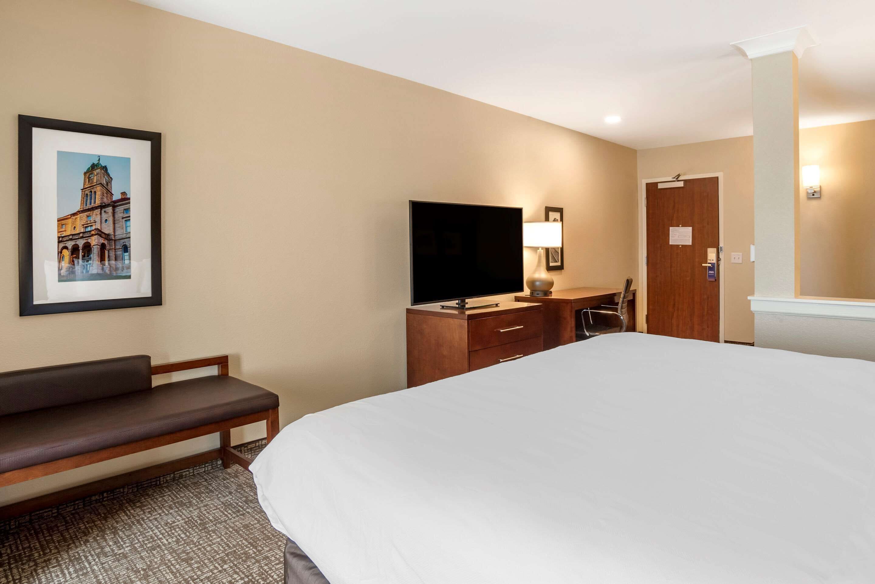 A hotel room with a large bed, a wooden bench, a desk with a flat-screen TV, and a framed picture on the wall.