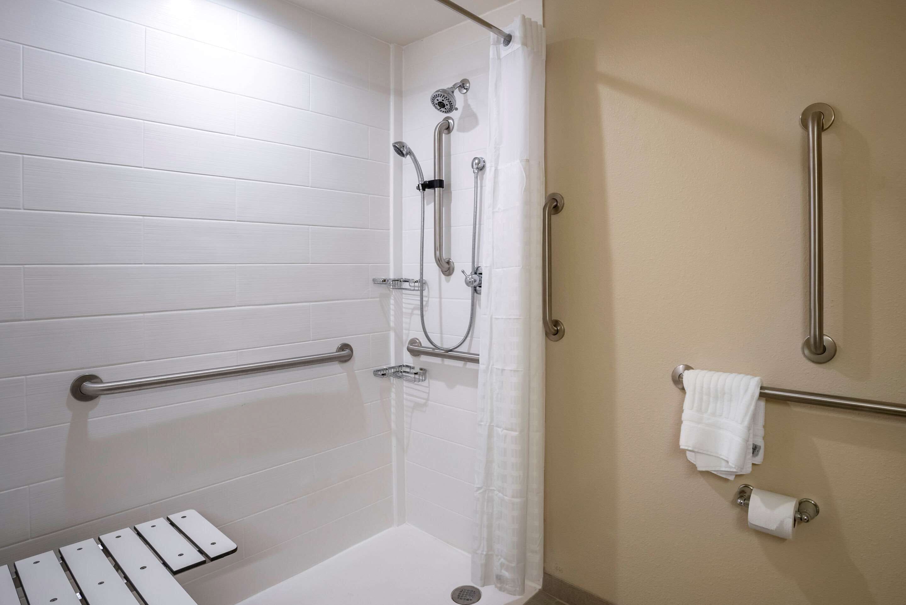 Un espace douche avec des murs recouverts de carreaux blancs, une barre d’appui, un pommeau de douche et un porte-serviettes.