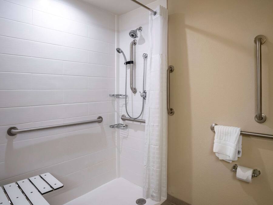 Un espace douche avec des murs recouverts de carreaux blancs, une barre d’appui, un pommeau de douche et un porte-serviettes.