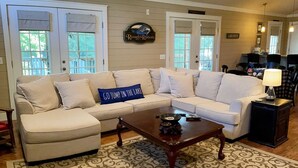 A living room with a beige sectional sofa, a coffee table, and a patterned rug. There are two doors leading outside, a sign above one door, and a lamp on a side table. - ROUGH RIVER LAKE Hot Tub, Pool, Grill, Fire Pit, Party Garage  (Falls of Rough)