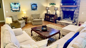 A living room with a stone fireplace, a flat-screen TV, and a large window. - ROUGH RIVER LAKE Hot Tub, Pool, Grill, Fire Pit, Party Garage  (Falls of Rough)