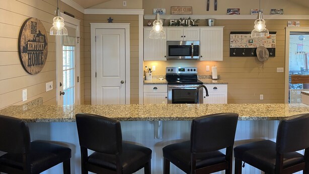 A kitchen with granite countertops, white cabinetry, a stainless steel oven, and a microwave. There are three black bar stools and a door with a sign above it. - ROUGH RIVER LAKE Hot Tub, Pool, Grill, Fire Pit, Party Garage  (Falls of Rough)