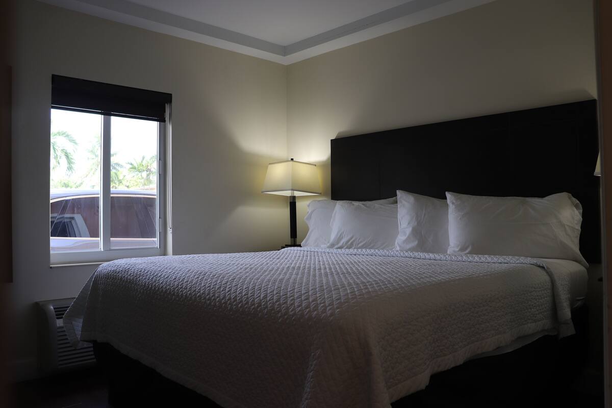 Una cama bien hecha con ropa blanca, una lámpara de noche y una ventana con persianas.