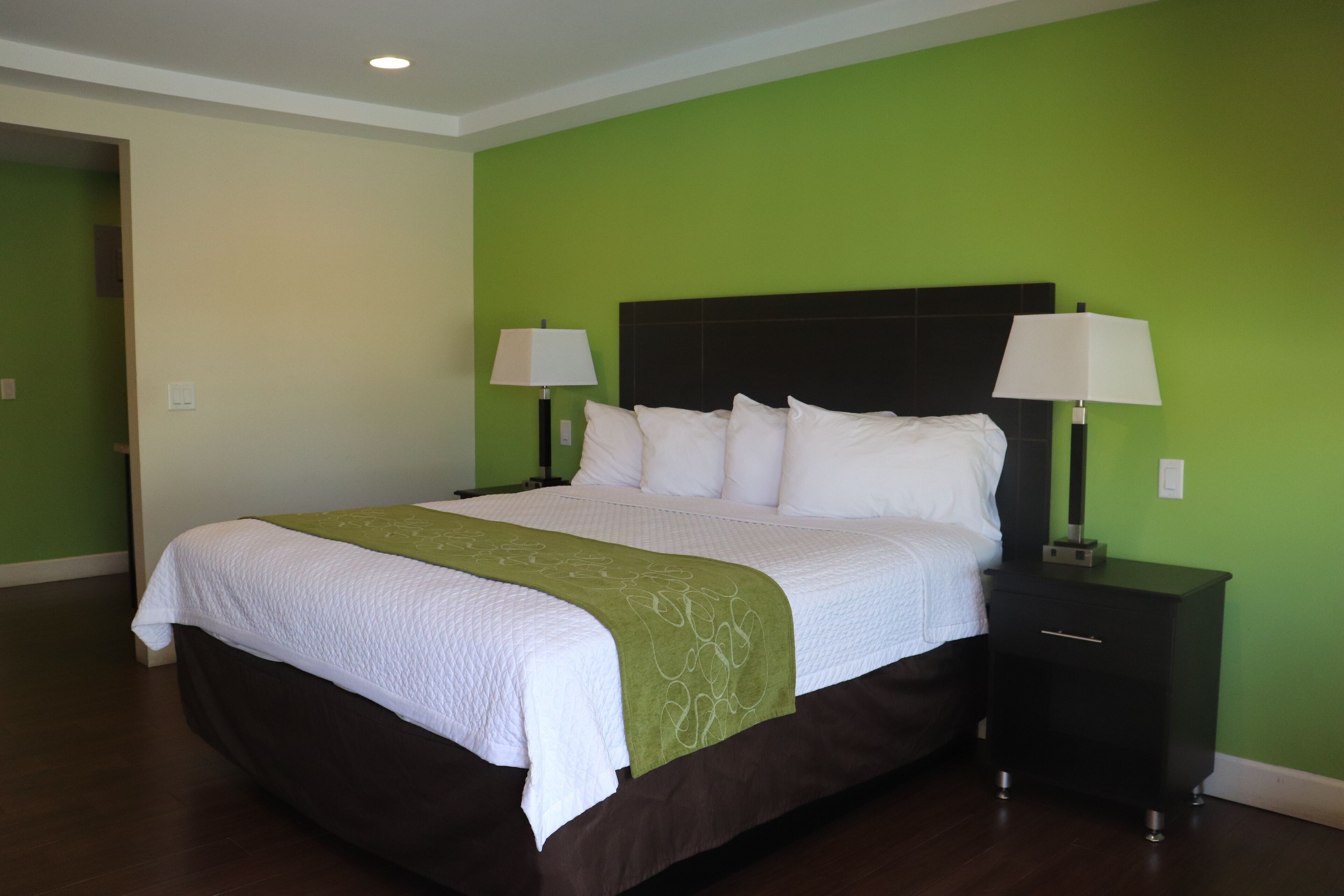 A neatly made bed with white linens and a green throw, flanked by two bedside tables with lamps, in a room with green walls and wooden flooring.