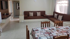 A living room with a brown sofa, a coffee table with a magazine cover, a TV on a wooden shelf, and a ceiling fan.