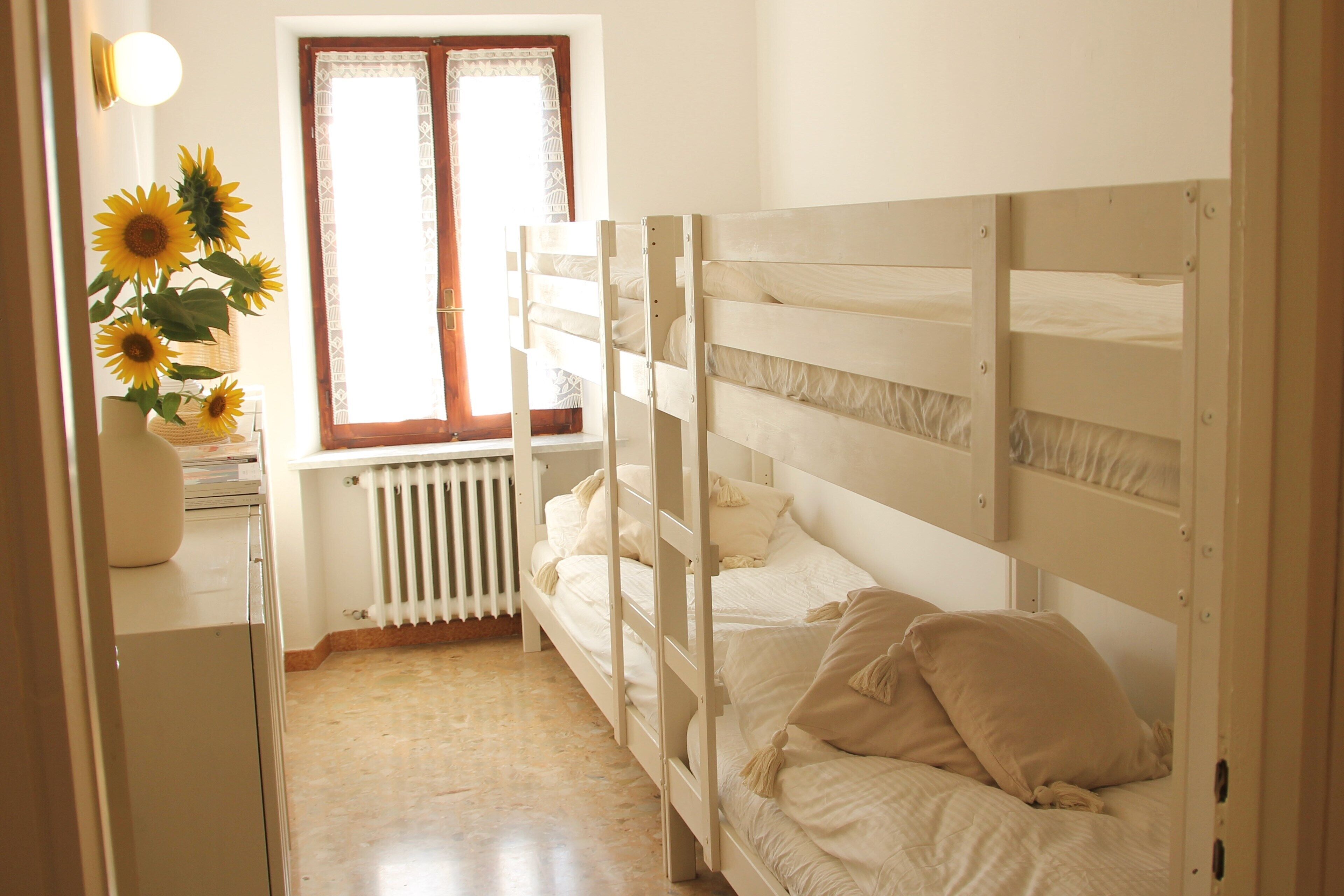 A bedroom with bunk beds, a radiator, a window with curtains, and a vase with sunflowers.