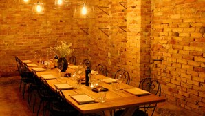 Un comedor con pared de ladrillo, mesa de madera, sillas de metal, lámparas colgantes y un jarrón con flores.