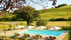 Un área de piscina con sillones de descanso y sombrillas, rodeada de un paisaje con césped y árboles.