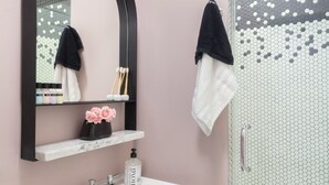 Baño con lavamanos de mármol, espejo, estante con artículos de aseo, ducha con azulejos hexagonales y toallas colgadas.
