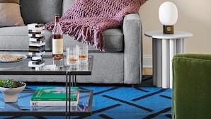 Un salón con un sofá, una mesa baja con libros y una botella, una mesita redonda con una lámpara, y un cuadro en la pared.