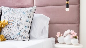 Un dormitorio con cabecera rosa, ropa de cama blanca, una mesita de noche pequeña con jarrones de flores y una lámpara colgante.