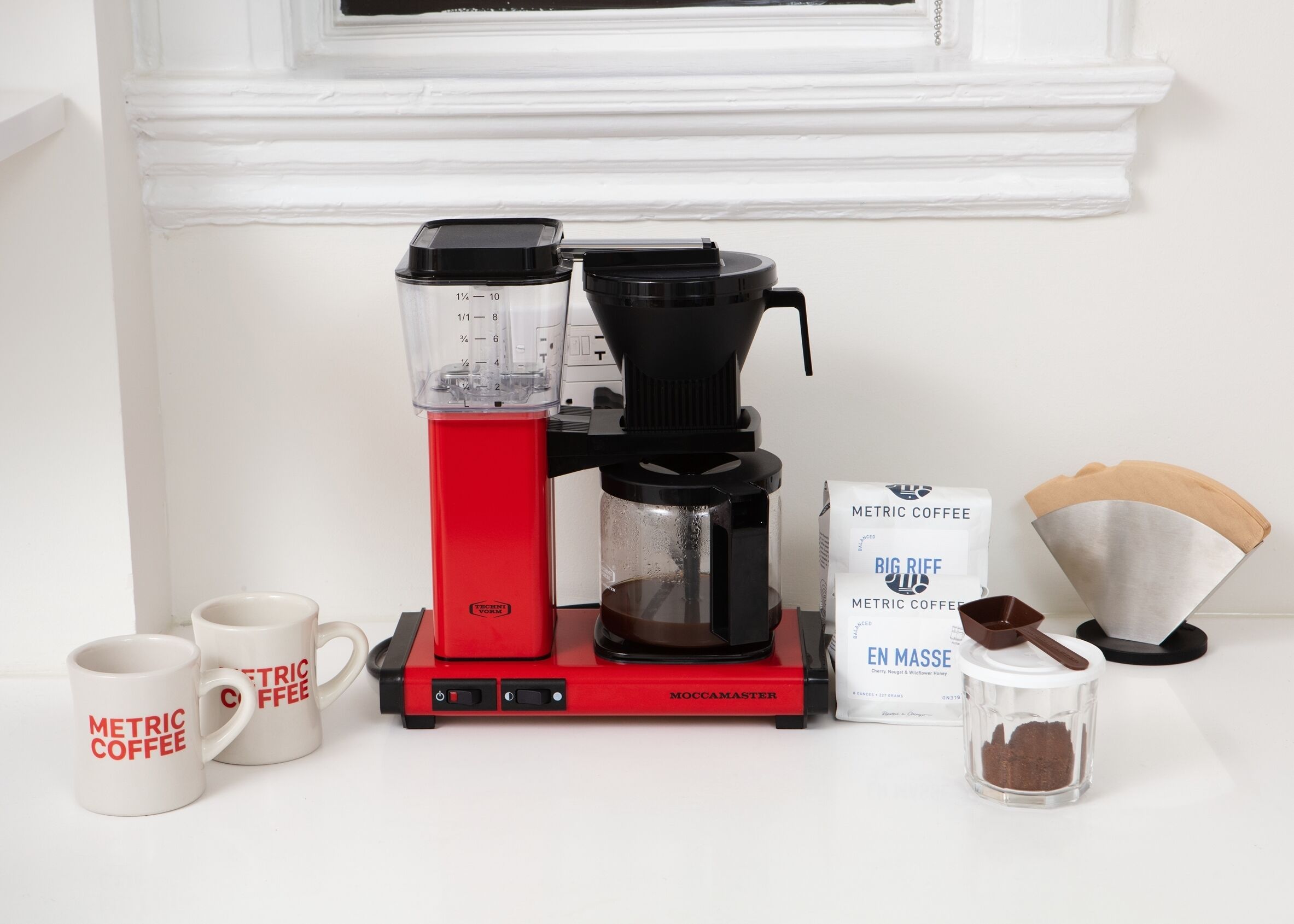 Een koffiezetapparaat met een rode basis, een koffiemolen, twee witte mokken met rode tekst en koffiekapsules op een witte aanrecht.