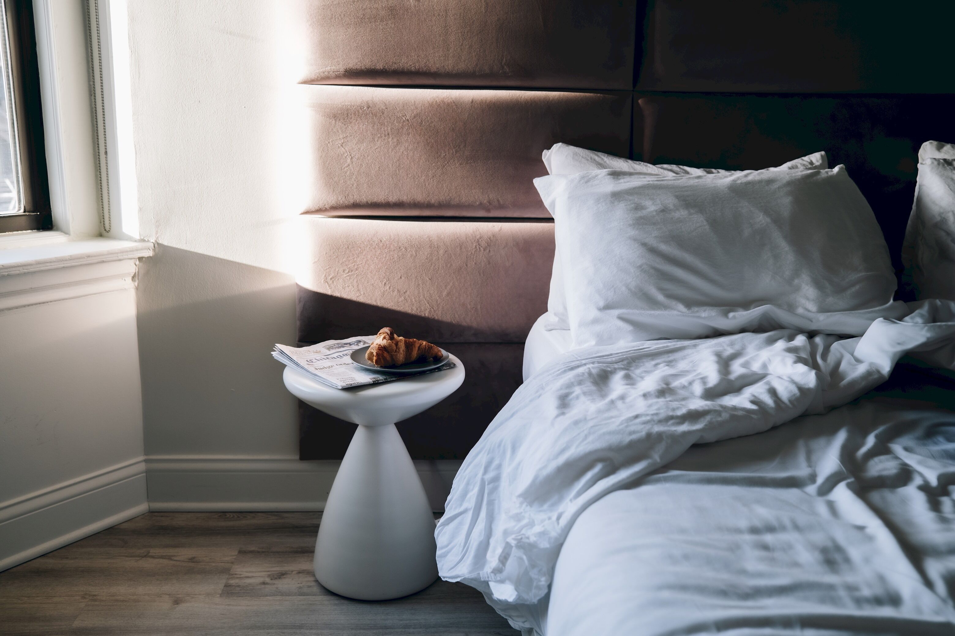 Een slaapkamer met een bed, kussens, een nachtkastje met een croissant, en een raam.